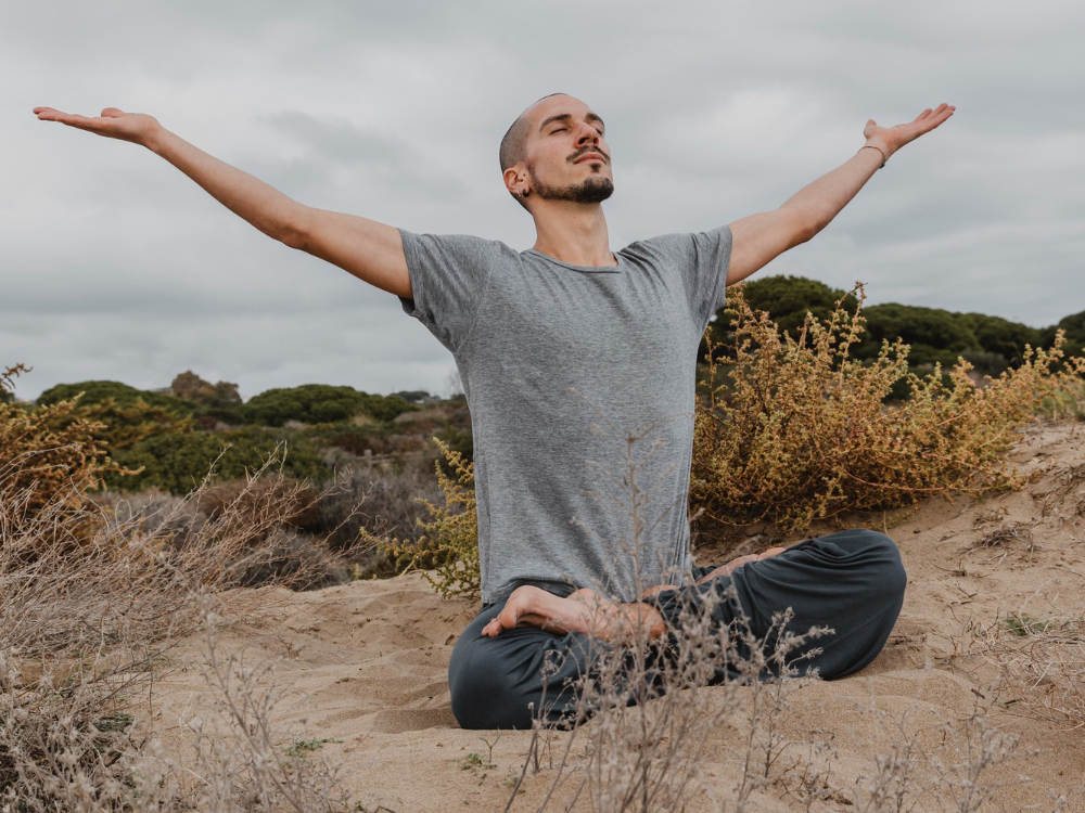 Person meditating outdoors in a gray shirt with arms outstretched.